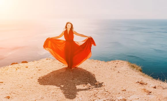 Side view a Young beautiful sensual woman in a red long dress posing on a rock high above the sea during sunrise. Girl on the nature on blue sky background. Fashion photo.