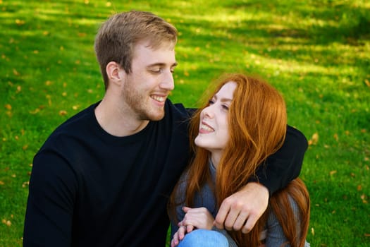 Happy young couple of Caucasian ethnicity man and woman in casual clothes sitting embracing in the park on green grass on a sunny day. Happy relationship between prana and a girl in nature