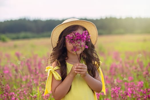 A beautiful playful little girl in a straw hat, with long dark hair, stands in a flowering field, in summer, holds a bouquet of burgundy viscaria flowers near her face. copy space. Close up