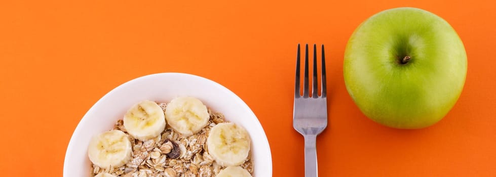 muesli with banana on orange background with apple. High quality photomuesli with banana in a white plate on an orange background, a fork and a green apple . Healthy food and diet concept