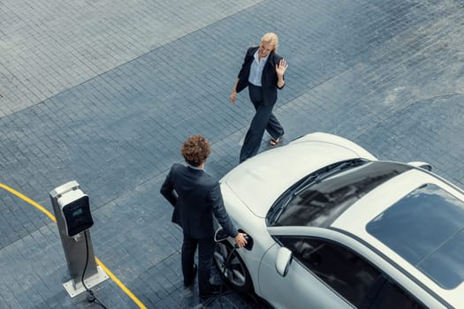 Aerial view progressive lifestyle of businesspeople with electric car connected to charging station at city center public parking car. Eco friendly rechargeable car powered by alternative clean energy