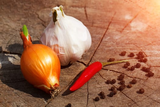 a head of garlic, onion and red hot pepper on a cracked wooden stump background close-up on a sunny summer day. the row is sprinkled with black pepper peas