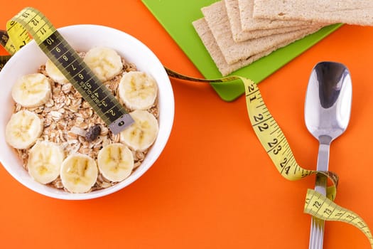 oatmeal with sliced banana and crispbread lie with spoon and yellow measuring tape on orange background, healthy food and diet concept