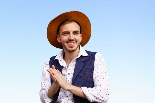 Portrait of a handsome young man in a hat against a blue sky. Caucasian guy with a goatee smiling happy summer outdoors sitting in a shirt and vest