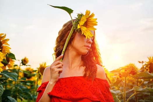 Cute young woman is holding sunflower in her hand while standing in field at sunset. Beautiful gentle girl of Caucasian ethnicity in red dress in the rays of setting sun. The concept of natural