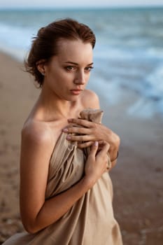 Lonely looking into the distance on the beach at sunset. Summer evening by the water. Beautiful young woman wrapped in a blanket. The girl on the seashore in the evening stands.