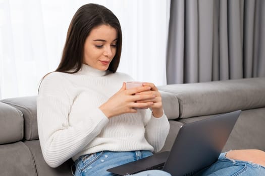 Beautiful brunette with long hair holding a cup watching video, having a pleasant conversation online using computer in cozy living room.