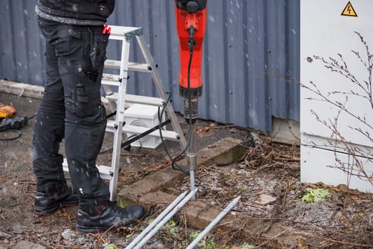 A worker installs a ground rod to ground a building. A worker in work clothes drives earth rods into the ground with a jackhammer to prevent short circuits