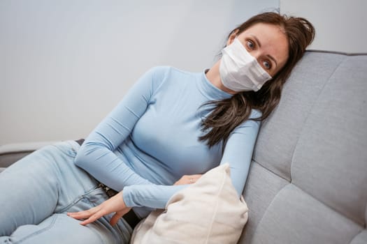 tired caucasian woman in casual clothes in a protective mask on the couch with a phone