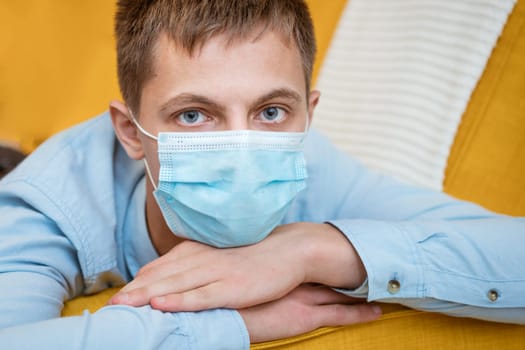 a young guy of Caucasian ethnicity in a shirt and a protective mask sits on the couch alone at home. Concept of being at home during global quarantine