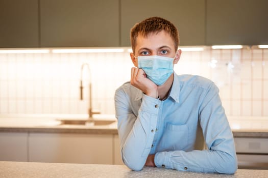 a young guy of Caucasian appearance sits in a light blue shirt and a medical mask in the kitchen alone at the table and ponders