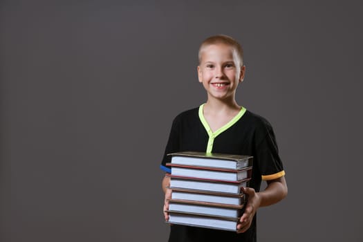 Cheerful caucasian boy in a black t-shirt holds a stack of books on a gray background. Education concept