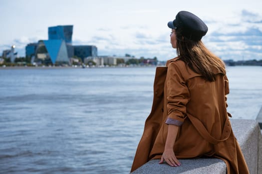 A sad lonely woman of European appearance in a jacket and a cap sits on the embankment of the river on a gloomy day, looks into the distance