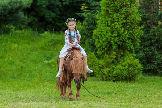 A little girl in the Ukrainian national costume rides a pony on the lawn