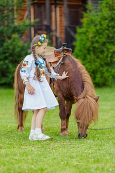 young girl in Ukrainian national dress strokes a pony that grazes on the lawn