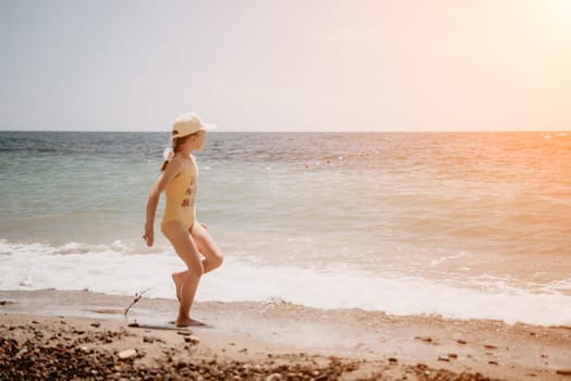 Cute little girl running along the seashore against a clear blue sea and rejoices in the rays of the summer sun. Beautiful girl in yellow swimsuit running and having fun on tropical beach