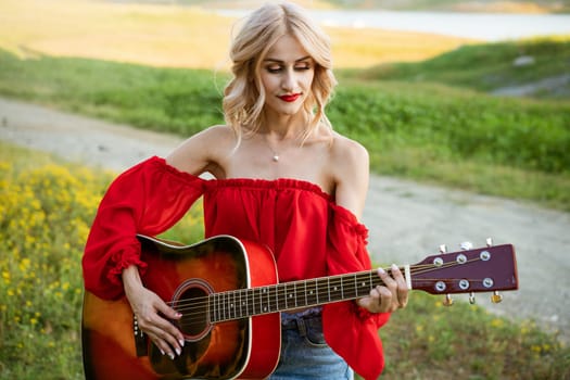 The woman in red is sitting on the green grass with a red guitar. Girl musician plays the guitar outdoors. Blonde Caucasian appearance