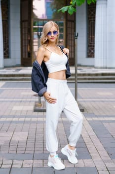 Young beautiful woman of Kakaz nationality poses in the afternoon on the street of the city in a white tracksuit and blue jeans. Confident blonde woman in sunglasses in summer