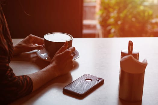 A young beautiful woman of Caucasian appearance sits at a table with a mug of coffee by the window in a cafe, looking to the side with a pensive look