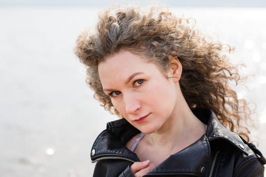 Portrait of a caucasian curly woman in a black leather jacket on the background of the bay looking at the camera