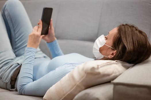 tired caucasian woman in casual clothes in a protective mask on the couch with a phone