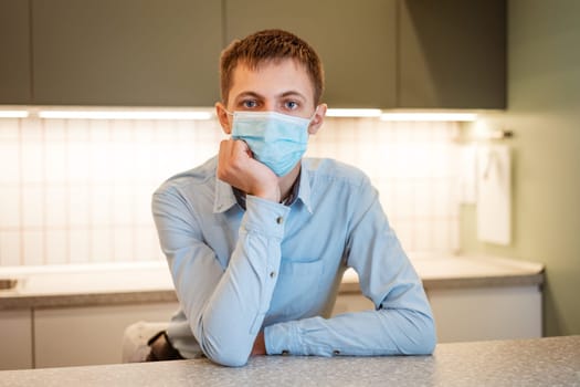 a young guy of Caucasian appearance sits in a light blue shirt and a medical mask in the kitchen alone at the table and ponders