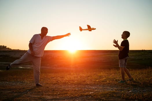 Soft focus of father and son playing toy airplane in meadow at sunset with happy emotions. Family, vacation and travel concept. At sunset in summer they launch an airplane against background sky
