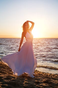 A beautiful young woman of Caucasian appearance with dyed hair posing on the seashore in the sun at sunset in a white dress in the wind. Romantic slender girl in nature enjoys relaxation