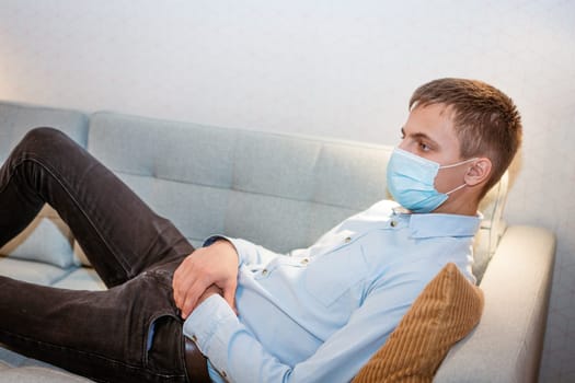 a young guy of Caucasian ethnicity in a shirt and a protective mask sits on the couch alone at home. Concept of being at home during global quarantine