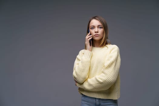 Young beautiful woman of Caucasian ethnicity talking on the phone in a yellow volumetric sweater on a gray background with blue eyes