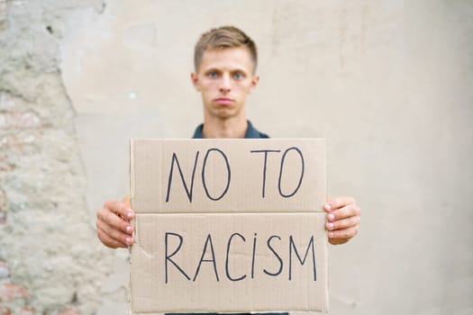 Caucasian guy came out to protest against racism with poster in his hands. Young man appearance stands against background of cement wall with cardboard in his hand on which it is written no to racism