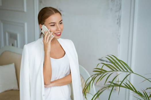 Successful young and beautiful woman of Caucasian ethnicity in a white jacket and dress talking on the phone, beautiful make-up hairstyle. Happy confident girl