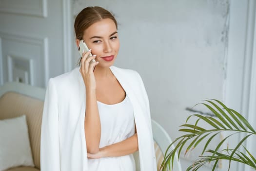 Successful young and beautiful woman of Caucasian ethnicity in a white jacket and dress talking on the phone, beautiful make-up hairstyle. Happy confident girl