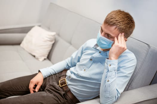 a young guy of European appearance, sitting comfortably on the couch in a medical mask. Concept to wait out the pandemic while sitting at home