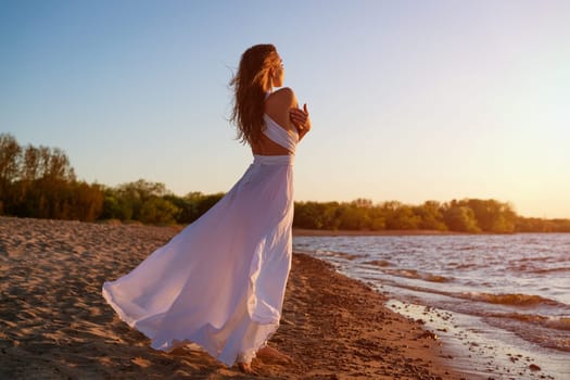A beautiful young woman of Caucasian appearance with dyed hair posing on the seashore in the sun at sunset in a white dress in the wind. Romantic slender girl in nature enjoys relaxation