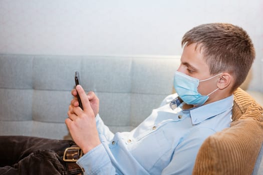 young caucasian man sitting on a sofa in casual clothes, wearing a protective medical mask with a phone in his hand