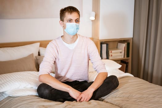 a young guy of European appearance, sitting comfortably on the couch in a medical mask. Concept to wait out the pandemic while sitting at home