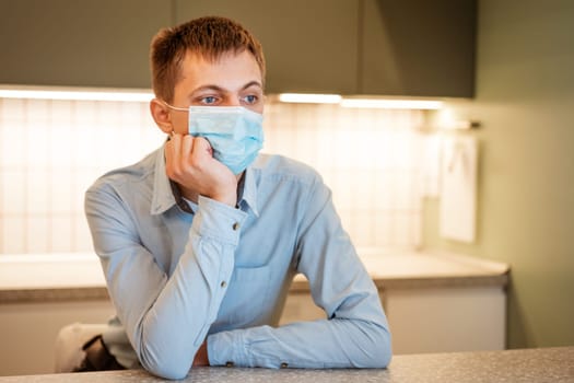 a young guy of Caucasian appearance sits in a light blue shirt and a medical mask in the kitchen alone at the table and ponders