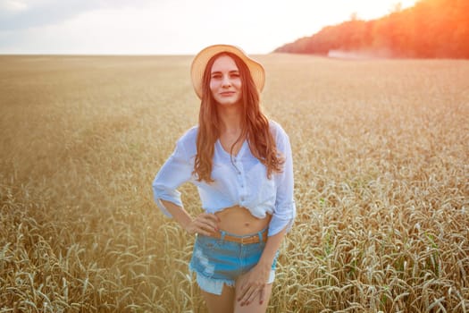 Happy girl sits in wheat field in straw hat. Cute young woman of Caucasian ethnicity in casual clothes enjoys ripe golden wheat in a field. Free woman concept