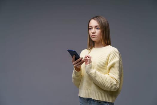 a young beautiful woman of Caucasian ethnicity writes sms on the phone, stands in a yellow sweater on a gray background, communicating via the Internet