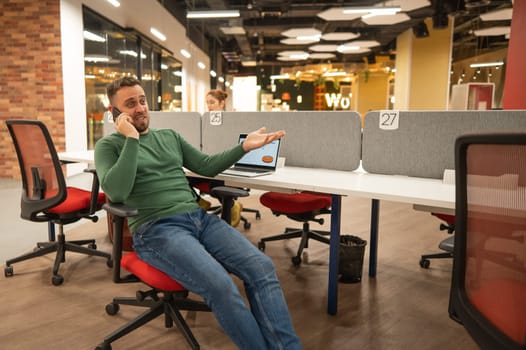 Caucasian bearded man talking on a smartphone while sitting at his desk in a modern coworking space