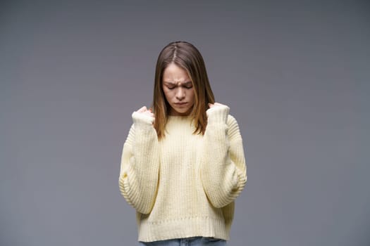 Portrait of a sad young woman of Caucasian ethnicity, clenches her fists in a yellow sweater on a gray background. Experiencing failure and solving problems