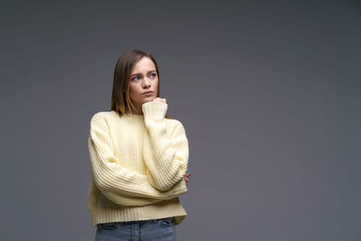 A young woman of Caucasian ethnicity, very upset in a yellow sweater on a gray background, holds her hands in fists. Emotional experiences and problem solving