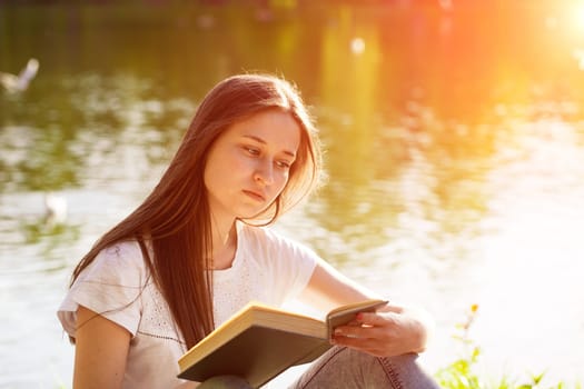 Young caucasian woman sitting by the lake in the park reading a book, sunny summer day