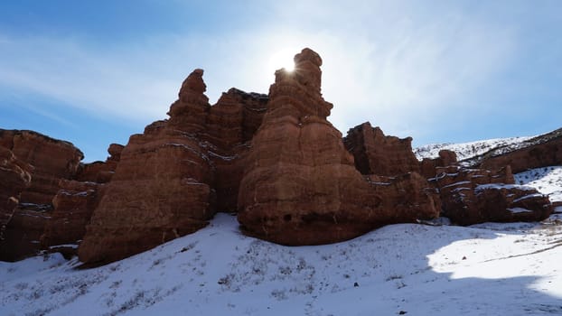 Grand Canyon Charyn. Orange cliffs and rocks. In places there are dry bushes and there is snow. The sun is shining brightly. White clouds. Faults and cracks in the canyon. The place looks like Mars