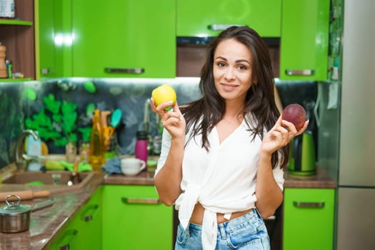 Young woman stands in kitchen holding fruits in her hands. Caucasian girl in a modern kitchen holds green apple and ripe peach in her hands. Chooses what to eat. Healthy nutrition for beautiful skin