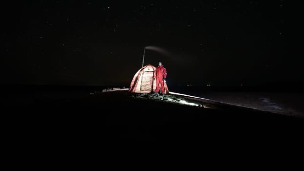 The guy is standing near a tent with a stove at night. Stars and constellations are visible. A lantern is burning in the tent, a pipe from a fresh camp stove is smoking. Bushes grow in places