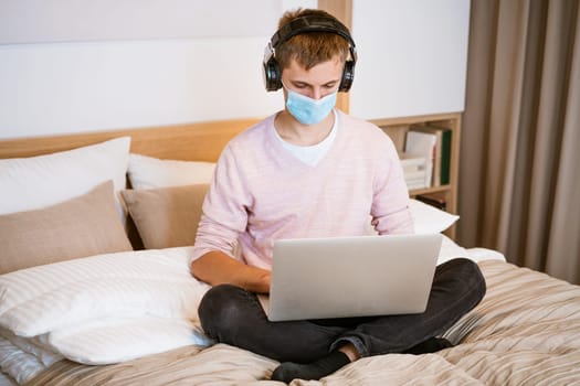 a young man of european appearance in casual clothes sits on a bed in a room at home wearing headphones and a protective mask at home with a laptop, the concept of remote work and education