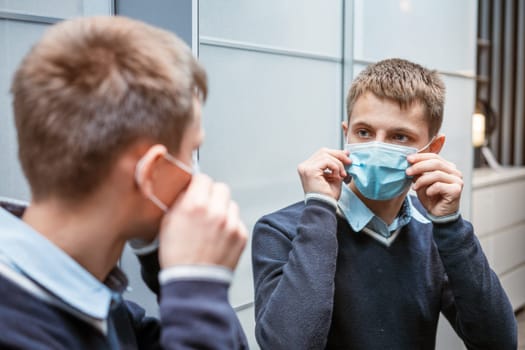 young caucasian man in a protective mask looks in the mirror, adjusts the medical mask against the virus. The concept of wave ipidemii