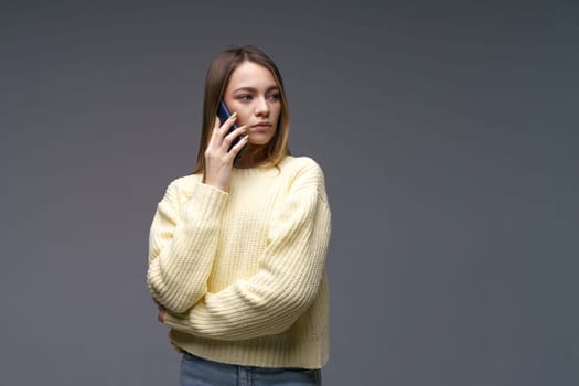 Young beautiful woman of Caucasian ethnicity talking on the phone in a yellow volumetric sweater on a gray background with blue eyes